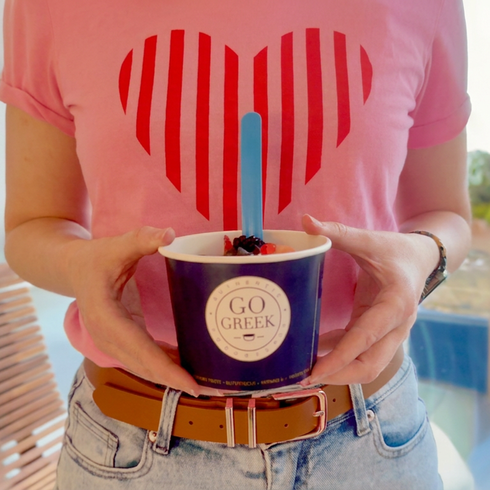Person in a pink shirt holds a Go Greek cup topped with berries and a blue spoon in a bright restaurant.