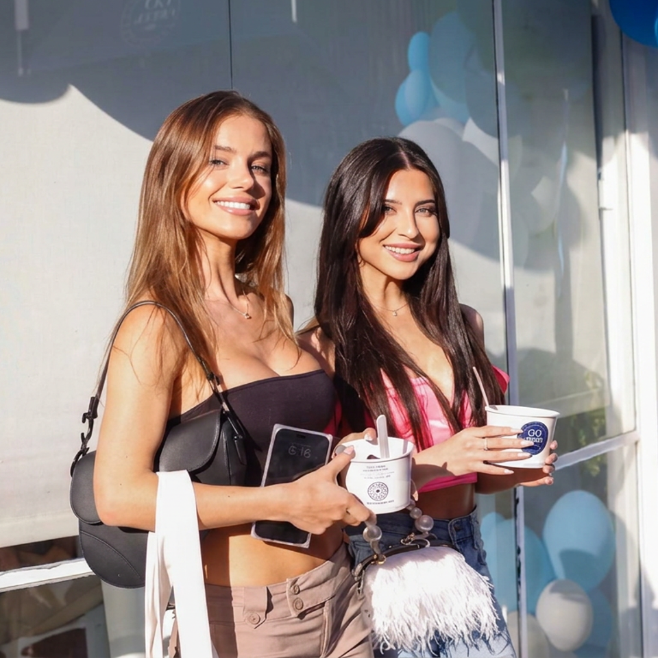 Two women smile outside a venue decorated with blue and white balloons while holding branded takeaway cups.