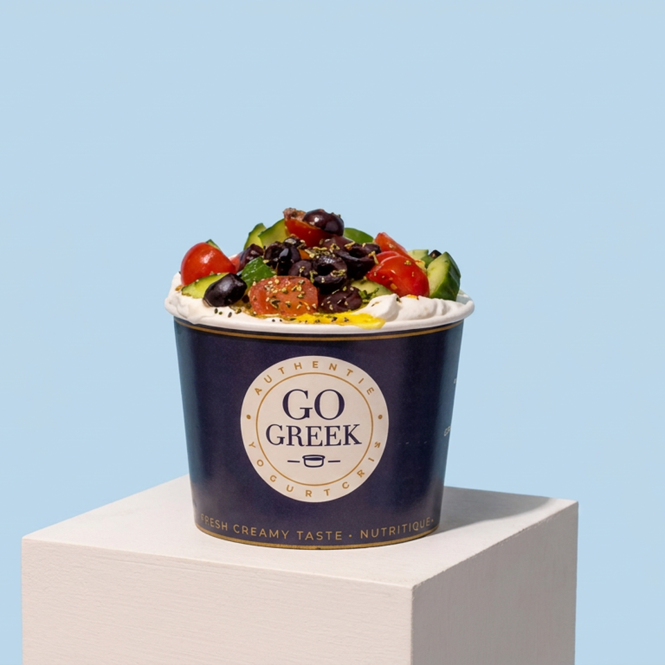 Greek yogurt bowl topped with tomatoes, cucumber, black olives, herbs, and olive oil in a branded cup on a pedestal.