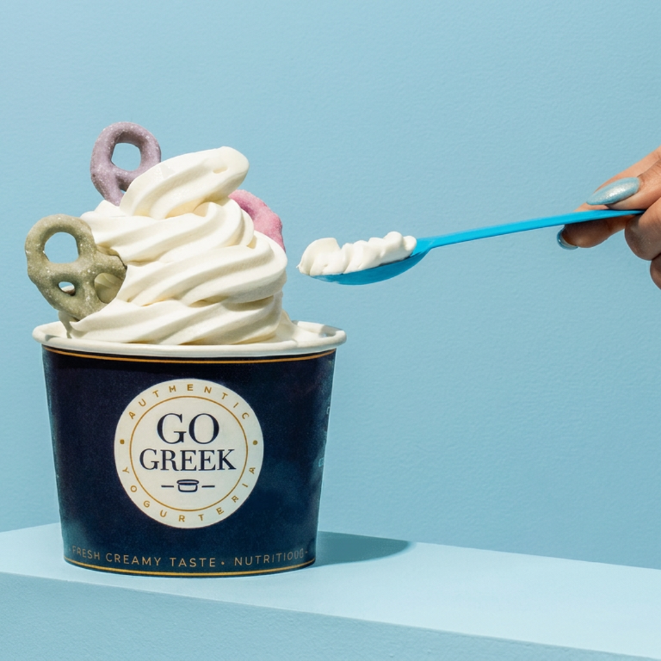Go Greek frozen yogurt topped with pastel candies, while a hand holds a blue spoonful beside it.