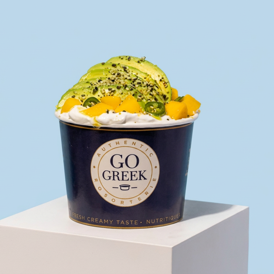 Go Greek yogurt cup topped with avocado slices, mango chunks, jalapeño, and sprinkled seeds on a white pedestal.