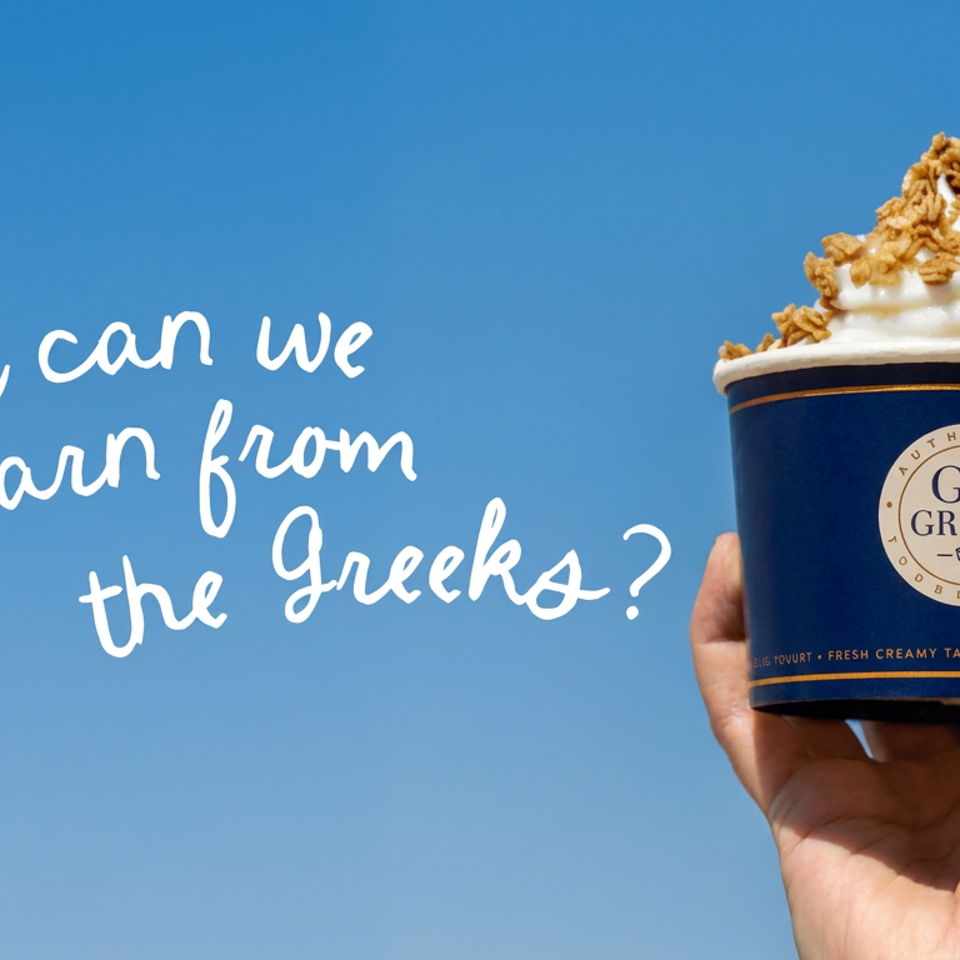 Hand holds a Go Greek cup topped with whipped cream and granola against a bright blue sky.