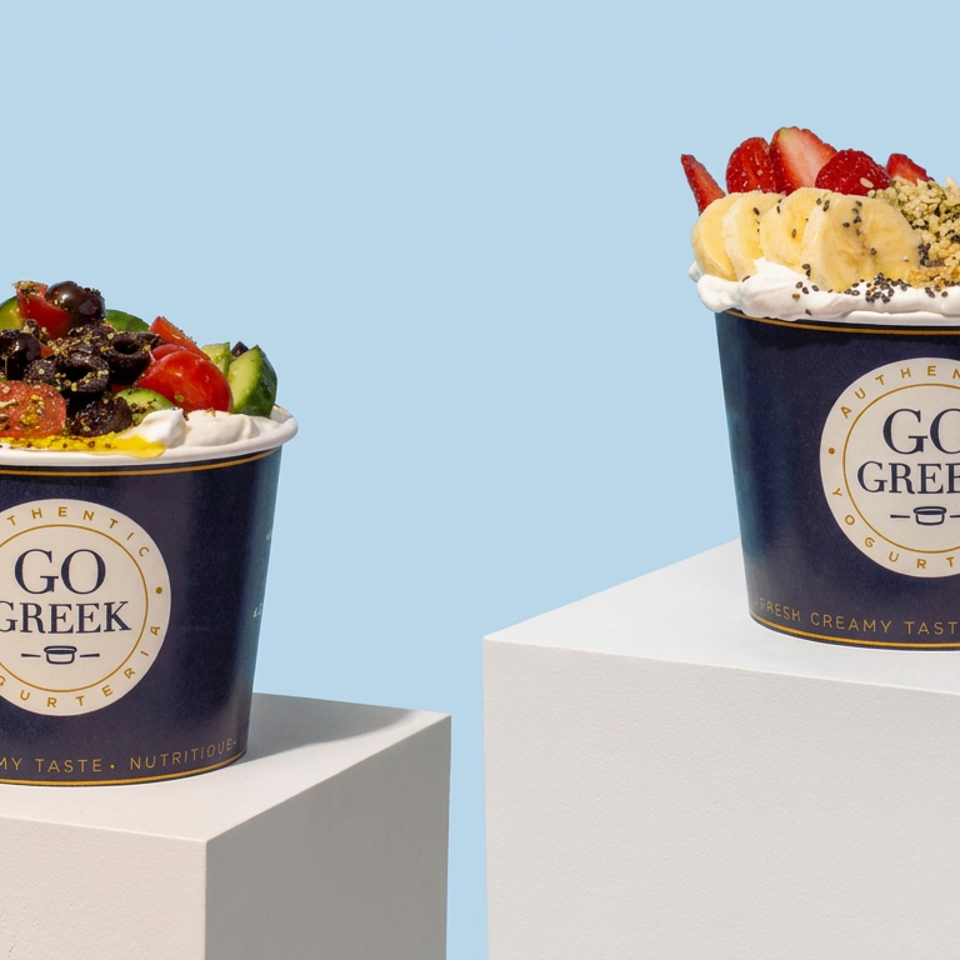 Two Go Greek yogurt cups topped with savory vegetables and olives, and fruit, granola, and berries, displayed on white pedestals.