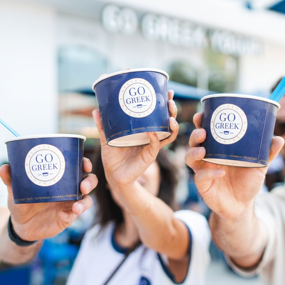 Three people hold blue Go Greek cups with straws toward the camera in a bright outdoor setting.