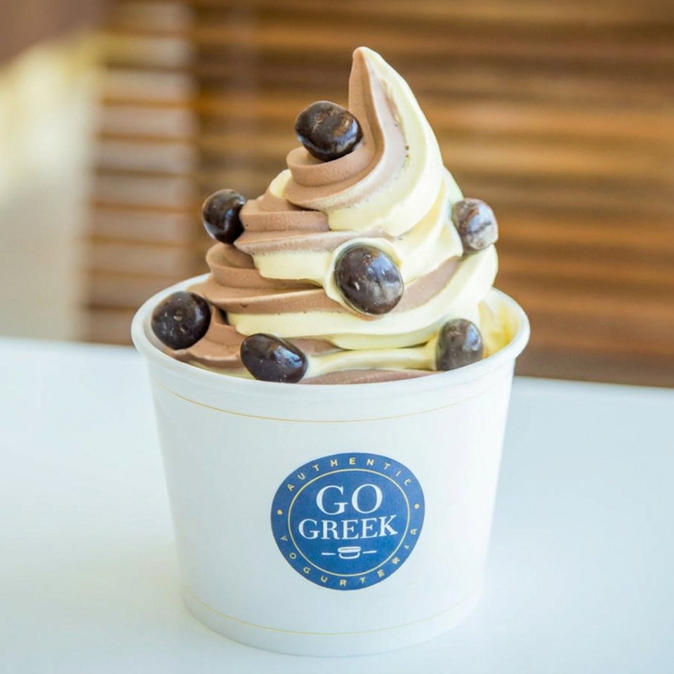 Swirled Greek yogurt topped with chocolate-coated berries in a Go Greek cup on a table.