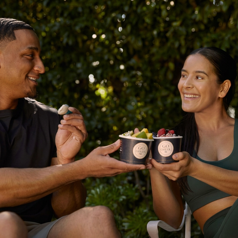 Two smiling people outdoors hold branded bowls topped with fresh fruit in a leafy garden setting.