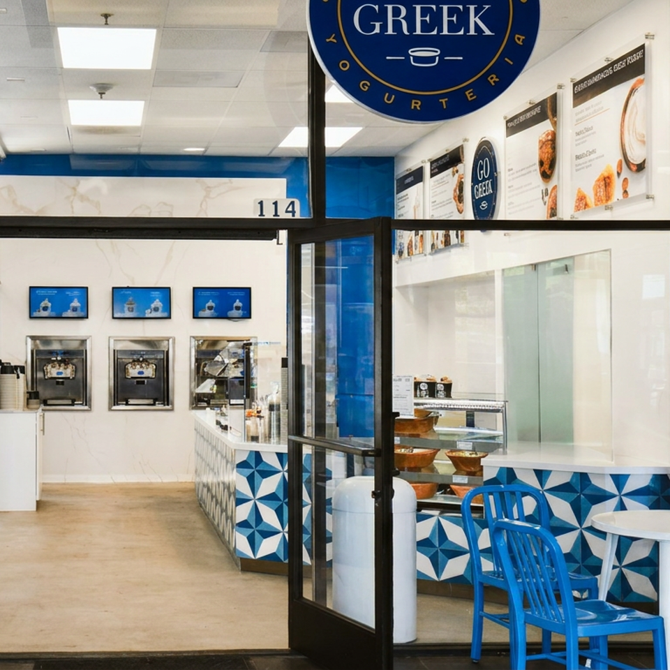 Bright Greek yogurt shop interior with blue chairs, patterned counter, self-serve machines, menu boards, and glass entrance.