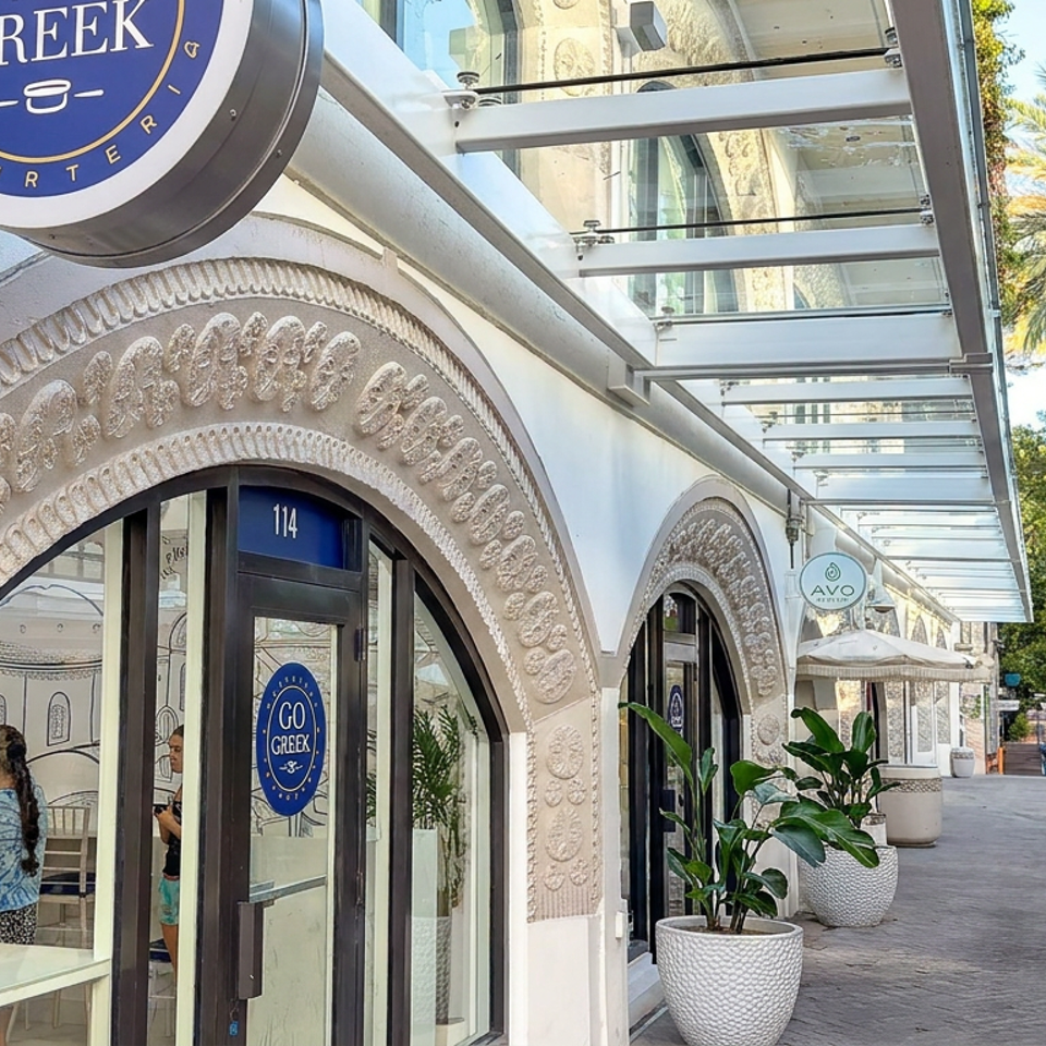 Go Greek yogurt shop entrance with ornate arched facade, blue signage, glass canopy, potted plants, and palm-lined walkway.